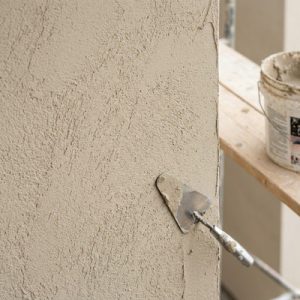 A metal trowel with fresh beige mortar sits against the textured, freshly layered stucco surface. In the blurred background, a wooden scaffold plank and a white bucket with plaster remnants are visible, emphasizing an active construction or masonry service site.