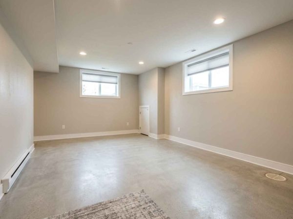An empty, wide-angle view of a clean, finished basement with waterproof polished concrete floors and light beige walls. The room features a wide, open floor plan with two high basement windows, recessed ceiling lights, white baseboards, and a subtle drain in the corner, giving it a bright and modern look.
