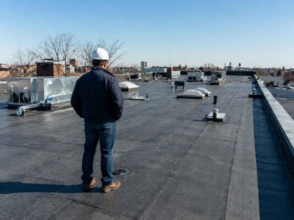 Professional roof inspector standing on a flat commercial roof conducting a roof inspection. The inspector is viewed from behind while examining the roofing surface, roof drains, vents, and rooftop equipment. The image shows a realistic flat roofing system under clear daylight with nearby residential buildings and trees in the background.