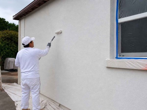 Professional worker applying stucco waterproofing coating to an exterior residential wall during the final finishing stage, creating a smooth, weather-resistant surface with protective coverings around windows and surrounding areas.
